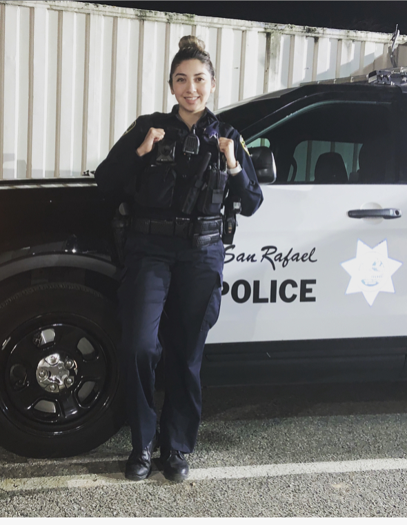 Officer Lorena Vega from the San Rafael Police Department smiles in uniform in front of her police vehicle.